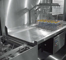 Outlet table beside a Hobart hood dishwasher with a wire rack of bottles under an overhead spray rail